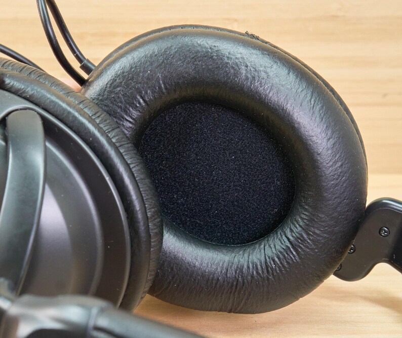 May include: Close-up of black over-ear headphones. The earcups are made of a soft, textured material, with a circular opening covered by a black, porous fabric. The headphones have a padded headband and are designed for comfort and sound isolation.