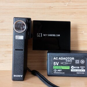 Peut inclure: Une caméra noire Sony GC1 Net-Sharing Cam, une boîte noire Net-Sharing Cam et un adaptateur secteur noir avec le texte "5V" et "JET". La caméra a un objectif et une sangle noire.