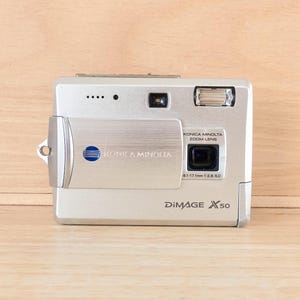 May include: A silver Konica Minolta DiMAGE X50 digital camera featuring a sliding lens cover, a built-in flash, and a zoom lens with 6.1-17.1mm 1:2.8-5.0 specifications. The front panel displays the Konica Minolta logo and the model name.