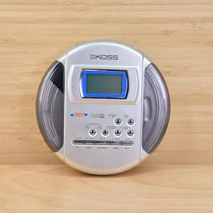 May include: Silver and black portable CD player with a blue-framed display screen. The device has a round design with the brand name "KOSS" at the top. It features control buttons and a clear plastic section on each side.