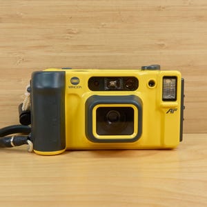 May include: A yellow Minolta AF film camera with a black grip and lens surround. The camera has a built-in flash and a wrist strap. The camera is on a wooden surface.
