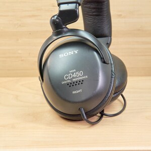 May include: Black Sony MDR-CD450 digital reference headphones. The headphones have a padded headband and earcups. The right earcup has the word "RIGHT" printed on it. The headphones are on a light brown wooden surface.