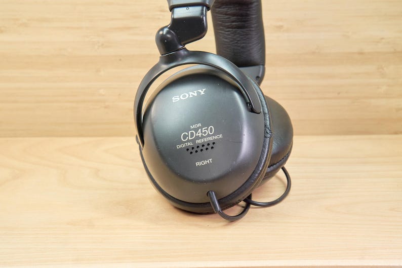 May include: Black Sony MDR-CD450 digital reference headphones. The headphones have a padded headband and earcups. The right earcup has the word "RIGHT" printed on it. The headphones are on a light brown wooden surface.