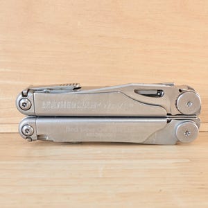 May include: A silver Leatherman Wave multi-tool in a closed, folded position. The metal handle features the engraved brand name Leatherman Wave and additional text on the lower section, secured with visible screws and a metal pocket clip.