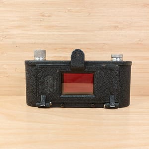 May include: A vintage black camera accessory with a textured surface, two silver knobs, and a red-tinted window. The accessory has a leather strap and metal clasps. The item is set against a light wood background.