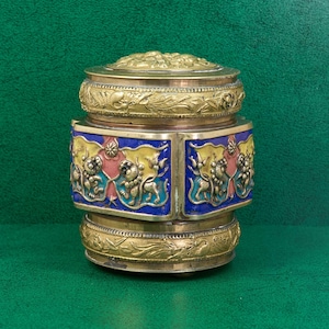 Peut inclure: Une boîte en laiton antique ornée d'un couvercle. La boîte présente des sculptures en relief détaillées et des accents d'émail colorés en bleu, jaune et rouge. Elle est probablement utilisée pour ranger de petits objets.