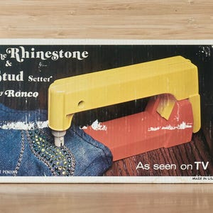 May include: A rectangular product box for The Rhinestone & Stud Setter by Ronco. The box features a yellow and red handheld tool applying rhinestones to blue denim fabric. Text includes The Rhinestone & Stud Setter, by Ronco, As seen on TV, and Patent Pending.
