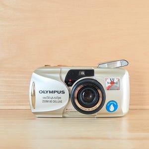 May include: A champagne-colored Olympus Stylus Epic Zoom 80 Deluxe camera with a retractable lens. The front features the text Olympus, Stylus Epic, Zoom 80 Deluxe, and Multi-AF. It includes a VF Technology sticker and a blue All Weather water drop icon.