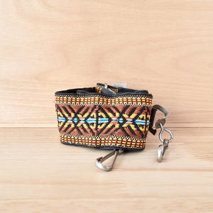 May include: A woven camera strap featuring a geometric pattern in shades of brown, yellow, and black with a thin horizontal blue stripe. The strap includes black fabric backing and two silver-toned metal swivel snap hooks at the ends.