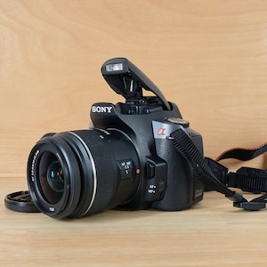 May include: A black Sony digital camera with an interchangeable lens and a pop-up flash unit. The camera body features the Sony logo on the front, an alpha symbol, and an attached black fabric neck strap with orange stitching details.
