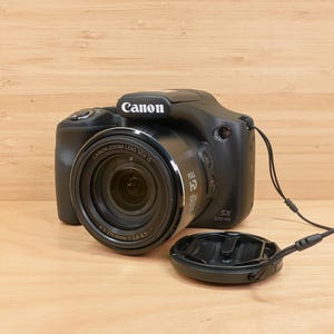 May include: Black Canon digital camera with a lens cap. The camera has a Canon Zoom Lens 50x IS. The lens is open and ready to take pictures. The camera is on a wooden surface.