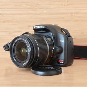 Op de afbeelding: Zwarte Canon EOS Rebel XSi DSLR-camera met zoomlens en lensdop. De camera heeft een zwarte band met rode rand. De lens is gemarkeerd met "Canon Zoom Lens EF-S 18-55mm 1:3.5-5.6 IS".