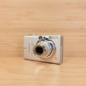 May include: Silver Canon PowerShot S410 digital camera with a retractable lens and flash. The camera has a silver body with black accents and a 4.0 megapixel sticker. The camera is on a wooden surface.