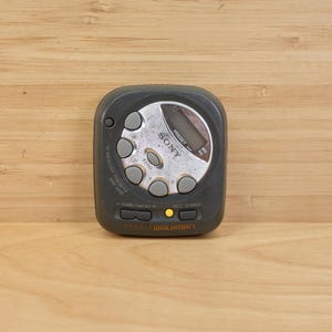 May include: A vintage Sony Walkman, a portable cassette player, in a dark grey casing. It features a silver control panel with buttons and a small digital display. The device is on a light brown wooden surface.