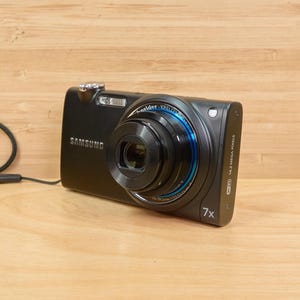 May include: A black Samsung digital camera with a 7x zoom lens. The camera has a blue ring around the lens and a silver button on the top.