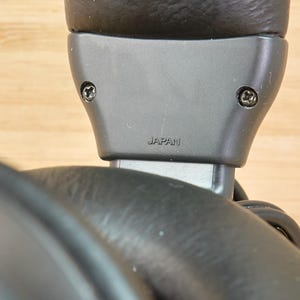 May include: Close-up of a black headphone component. The gray plastic piece has two screws and the word "JAPAN" printed on it. The headphone's ear cup is visible in the foreground, with a wooden surface in the background.