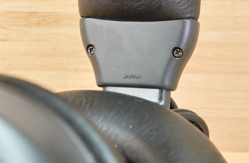 May include: Close-up of a black headphone component. The gray plastic piece has two screws and the word "JAPAN" printed on it. The headphone's ear cup is visible in the foreground, with a wooden surface in the background.