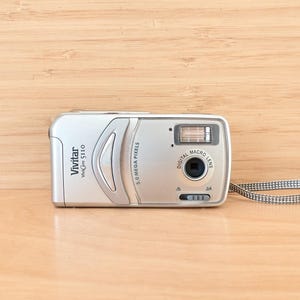 May include: Silver Vivitar Vivicam 5110 digital camera with a 5.0 megapixel lens. The camera has a built-in flash and a macro lens. A grey and white patterned strap is attached to the side of the camera.