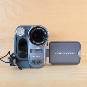 May include: A grey Sony Handycam video camera with an open side screen. The lens features 20x optical zoom and 990x digital zoom text. A circular light is positioned above the lens, and a black wrist strap with a lens cap is attached to the side.