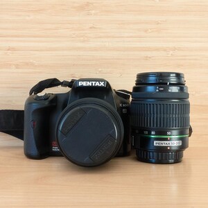 May include: Black Pentax K100D DSLR camera with a lens and lens cap. The lens is a Pentax 50-200mm. The camera has a black strap attached. The background is a light brown wood grain.