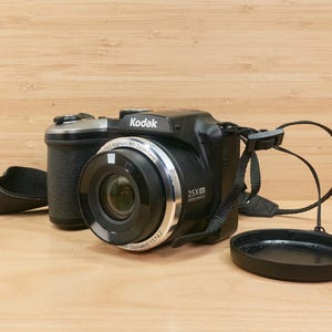 May include: A black Kodak digital camera with a silver lens ring and a 25X zoom lens. The camera has a black strap and lens cap. The camera is on a light brown wooden surface.