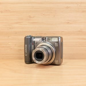 May include: A silver Canon PowerShot A590 IS digital camera with a zoom lens. The camera has a built-in flash and a 8.0 MP resolution. The camera is on a light brown wooden surface.