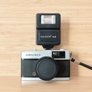 May include: An Olympus Trip 35 camera with a black and silver body, a black lens cap, and a black flash unit on top. The flash is labeled "SOLIGOR 14S". A black wrist strap is attached to the camera.