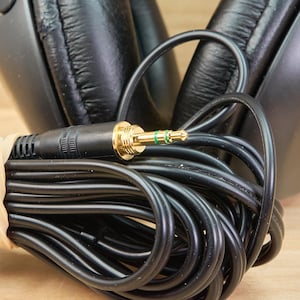 May include: Black over-ear headphones with a coiled black cable and a gold-colored 3.5 mm audio jack. The cable is secured with a beige rubber band. The headphones are on a light brown wooden surface.