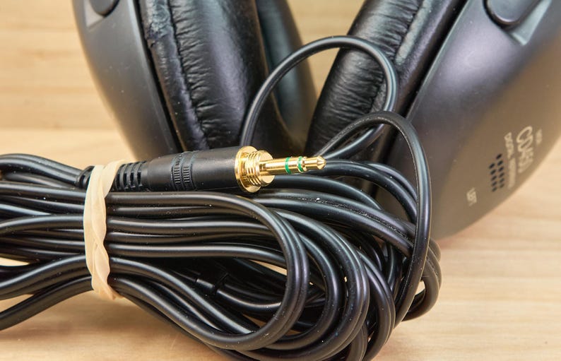 May include: Black over-ear headphones with a coiled black cable and a gold-colored 3.5 mm audio jack. The cable is secured with a beige rubber band. The headphones are on a light brown wooden surface.