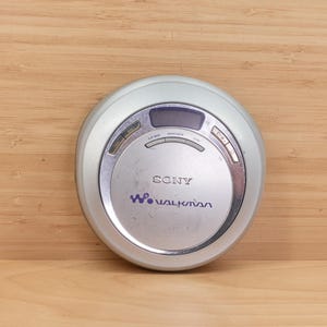 May include: A silver Sony Walkman CD player, circular in shape. It features a digital display and control buttons. The Sony and Walkman logos are visible. The device is set against a wooden background.