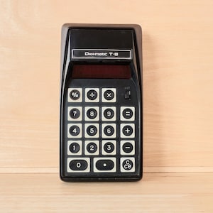Peut inclure: Une calculatrice électronique noire Digi-matic T-8 dotée d'un écran sombre, d'une grille de touches carrées blanches avec des chiffres et des symboles mathématiques noirs, et d'un petit interrupteur noir situé sur le côté droit de l'appareil.