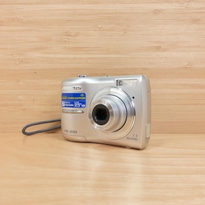 May include: Silver Olympus FE-210 digital camera with a retractable lens and a built-in flash. The camera has a blue sticker with text and a grey wrist strap. The camera is on a light brown wooden surface.