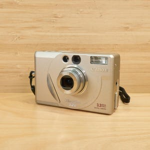 以下が含まれることがあります： 格納式レンズ、フラッシュ、リストストラップを備えた、シルバーのCanon PowerShot S20デジタルカメラ。 カメラは3.3メガピクセルの解像度で、明るい茶色の木製の背景に設定されています。