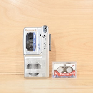May include: A silver Panasonic RN-205 microcassette recorder featuring a built-in speaker, a microphone port, and the text AstraZeneca printed on the side. Beside the recorder is a clear plastic case containing a Panasonic MC-60 microcassette tape.