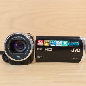 以下が含まれることがあります： 40倍光学ズームのコニカミノルタHDレンズを搭載した黒いJVCフルHDビデオカメラ。このデバイスには、3.0インチディスプレイ、Wi-Fi接続、AVCHD録画機能が含まれており、側面にはさまざまな技術仕様ラベルが印刷されています。