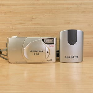 May include: A silver Olympus D-380 digital camera with a flash and the text "2.0 Megapixels" next to a silver SanDisk memory card reader. The camera has a strap and is on a wooden surface.