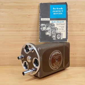 Könnte beinhalten: Eine braune Vintage Bell & Howell Auto Master Filmkamera mit silberfarbenen Akzenten. Die Kamera wird von einem Büchlein mit dem Titel "How to Make Perfect Movies" begleitet. Das Büchlein hat einen blau-schwarzen Einband.