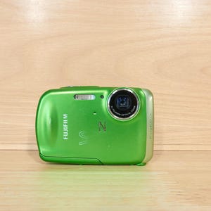 Könnte beinhalten: Eine leuchtend grüne Fujifilm FinePix Digitalkamera mit metallischem Finish. Die Vorderseite zeigt ein kreisförmiges Objektiv, einen kleinen Blitz und das Fujifilm-Logo in Weiß. Der Buchstabe Z ist auf das Gehäuse geprägt und der Name FinePix ist sichtbar.