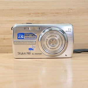 May include: Silver Olympus Stylus 760 all-weather digital camera with a 7.1 megapixel resolution. The camera has a zoom lens and a gray wrist strap. The camera has the text "DUAL IMAGE IS STABILIZATION" and "Stylus 760 ALL-WEATHER".