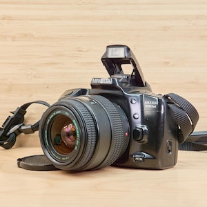 May include: Black Minolta Maxxum 400si camera with a lens and flash. The camera has a black strap and is sitting on a light brown wooden surface. The lens cap is off.