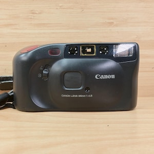 May include: A black Canon 35mm f/4.5 point-and-shoot film camera with a red button and a built-in flash. The camera has a leather strap and is on a light wood surface.