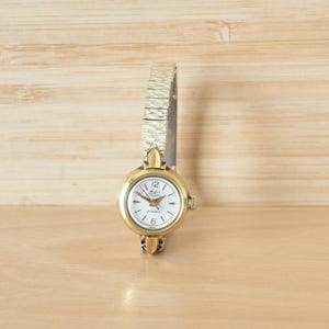 May include: A vintage gold-toned wristwatch with a white face and gold hands. The watch has a textured gold band and a small, round face with the numbers 12 and 6 visible. The watch is displayed on a light wooden surface.