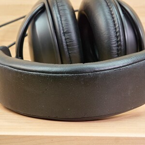May include: Black over-ear headphones with a padded headband and earcups. The headphones have a black cord that is bundled with a rubber band. The headphones are on a light brown wooden surface.