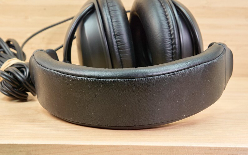 May include: Black over-ear headphones with a padded headband and earcups. The headphones have a black cord that is bundled with a rubber band. The headphones are on a light brown wooden surface.