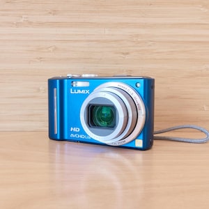 Peut inclure: Un appareil photo numérique compact bleu vif doté d'un objectif Lumix avec Power O.I.S. et un grand angle de 25 mm. L'avant de l'appareil affiche le logo Lumix, la mention HD et le texte AVCHD Lite, accompagné d'une dragonne grise.