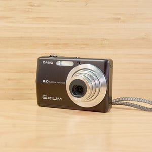 May include: A black Casio Exilim digital camera with a silver lens and a grey wrist strap. The camera has the text "CASIO", "6.0 MEGA PIXELS", and "EXILIM" printed on it. The camera is on a wooden surface.