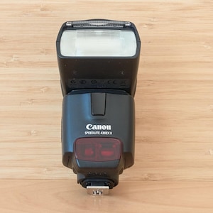 May include: A black Canon Speedlite 430EX II camera flash unit featuring a rectangular white flash head, a central control body with the brand name and model printed in white, and a red sensor window positioned above the metal mounting foot.