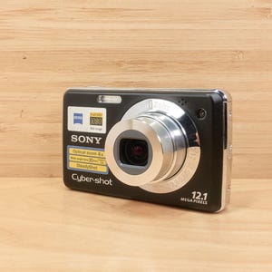 May include: Black Sony Cyber-shot digital camera with a silver lens and a 12.1 megapixel resolution. The camera has a flash and several stickers with technical information. The camera is on a wooden surface.
