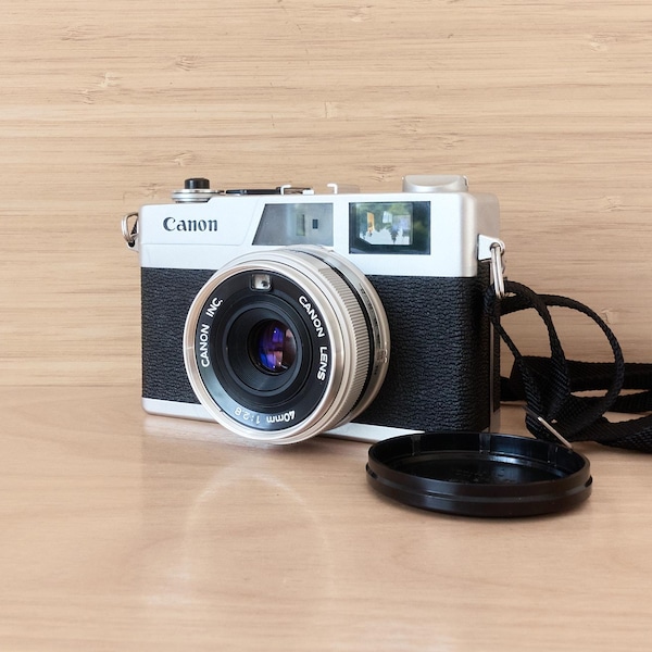 以下が含まれることがあります： 40mm f/1.9レンズを搭載したシルバーとブラックのキヤノン製レンジファインダーカメラ。カメラにはテクスチャ加工された黒いグリップ、キヤノンのロゴが入ったシルバーのトッププレート、ファインダー窓、黒いレンズキャップ、黒いネックストラップが付属しています。