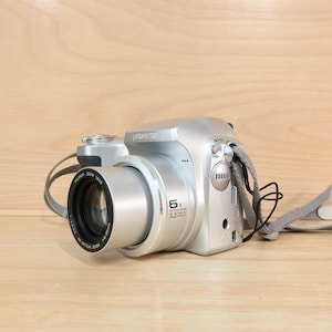 Peut inclure: Un appareil photo numérique argenté doté d'un objectif proéminent avec l'inscription 6x optical zoom et 3.2 mega pixels. Le boîtier comprend un déclencheur, une molette de sélection et une sangle de cou en tissu gris fixée sur le côté.